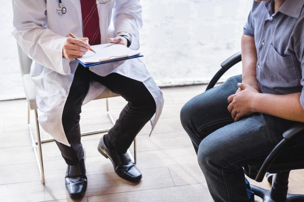 Ein Arzt in weißem Kittel sitzt mit einem Klemmbrett und Stift vor einem Patienten, der mit gefalteten Händen auf einem Stuhl sitzt.