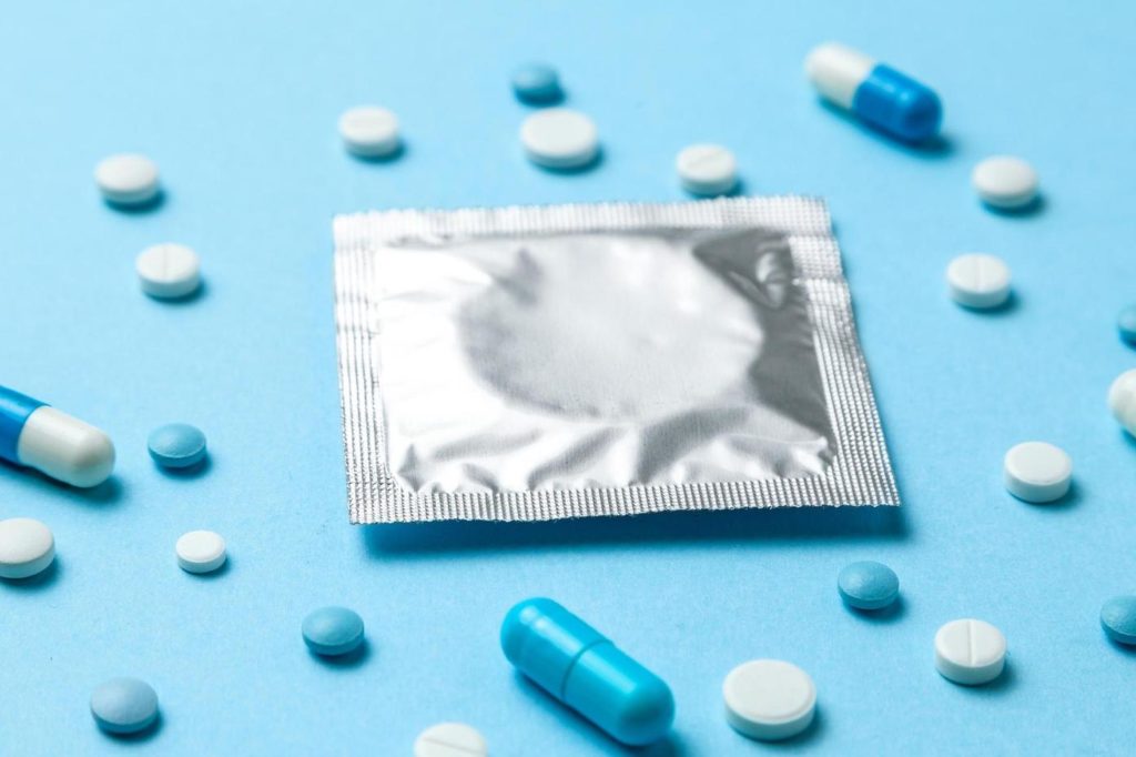 Verschiedene blaue und weiße Viagratabletten sowie eine verschlossene silberne Kondomverpackung liegen verstreut auf einem blauen Hintergrund.