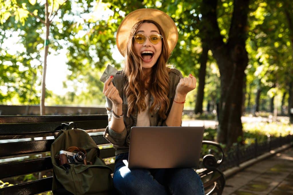 Eine junge Frau mit Hut und Sonnenbrille sitzt im Park auf einer Bank mit einem Laptop, hält eine Kreditkarte und jubelt vor Freude über ihren Kauf.
