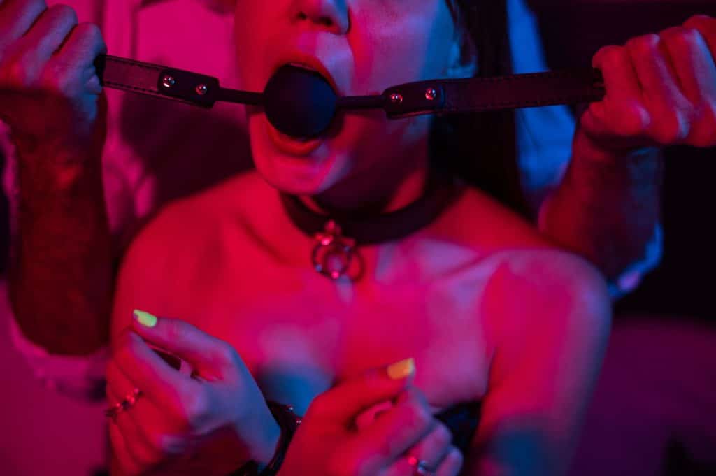 Eine Frau mit Knebel und Halsband in einer BDSM-Szene mit violetter Beleuchtung.